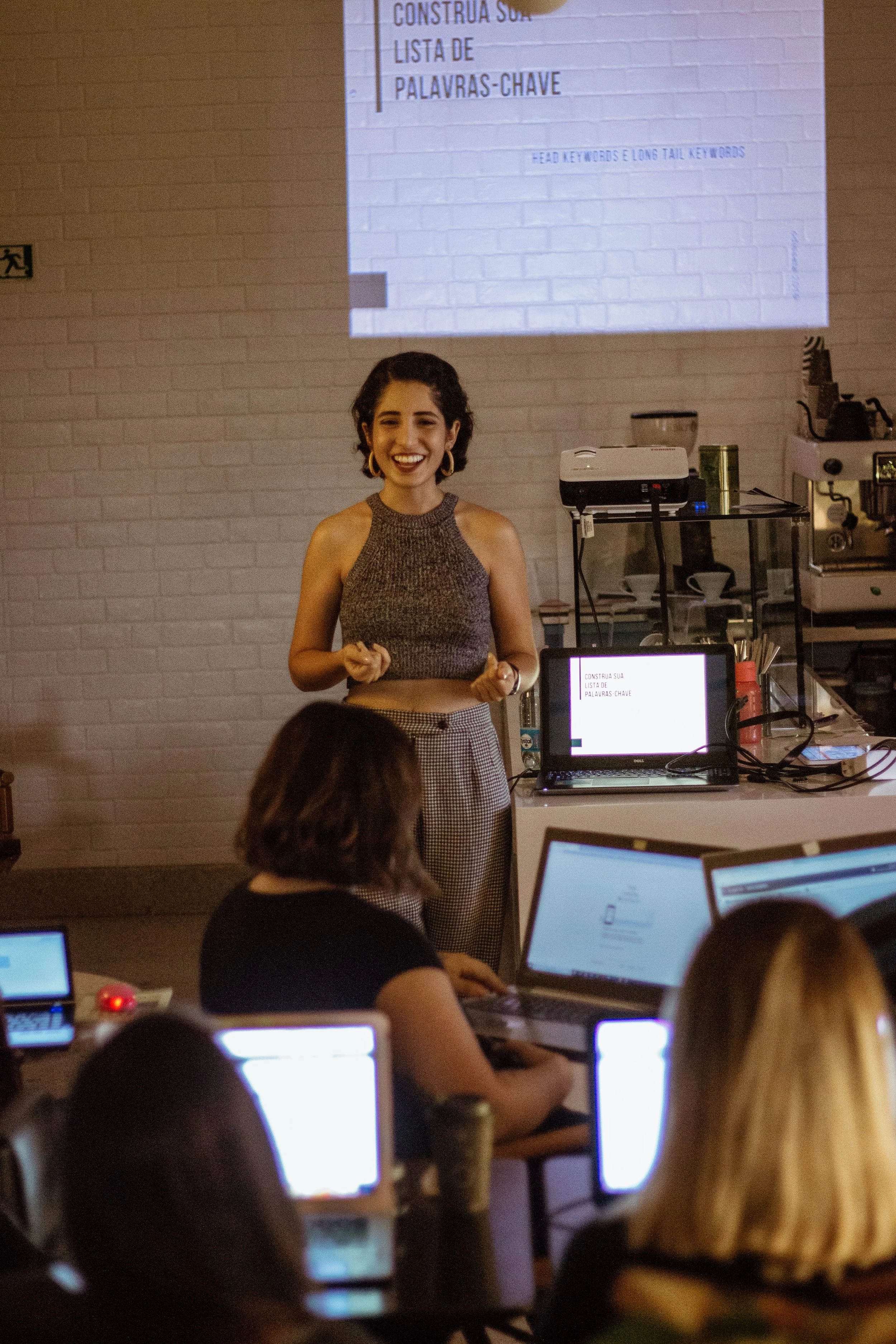 Young woman giving a presentation to an audience of people working on laptops, with a slide projected behind her that has Portuguese text about keywords.