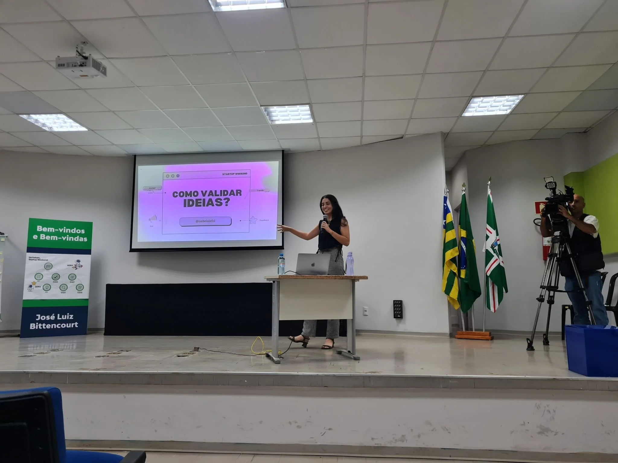A woman giving a presentation in a conference room with a large screen behind her that displays a slide titled 'Como Validar Ideias?' She is standing behind a desk with a laptop, water bottles, and a microphone, and is pointing towards the screen. A 