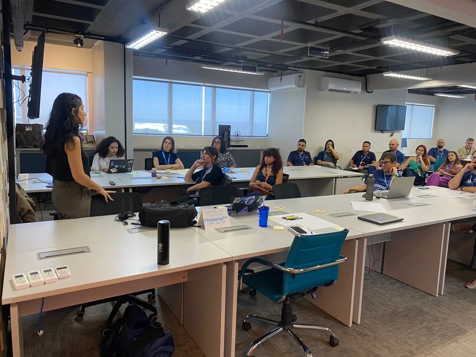 A woman stands in front of a group of people seated at office-style tables in a conference room. The room has large windows, ceiling lights, and wall-mounted screens.