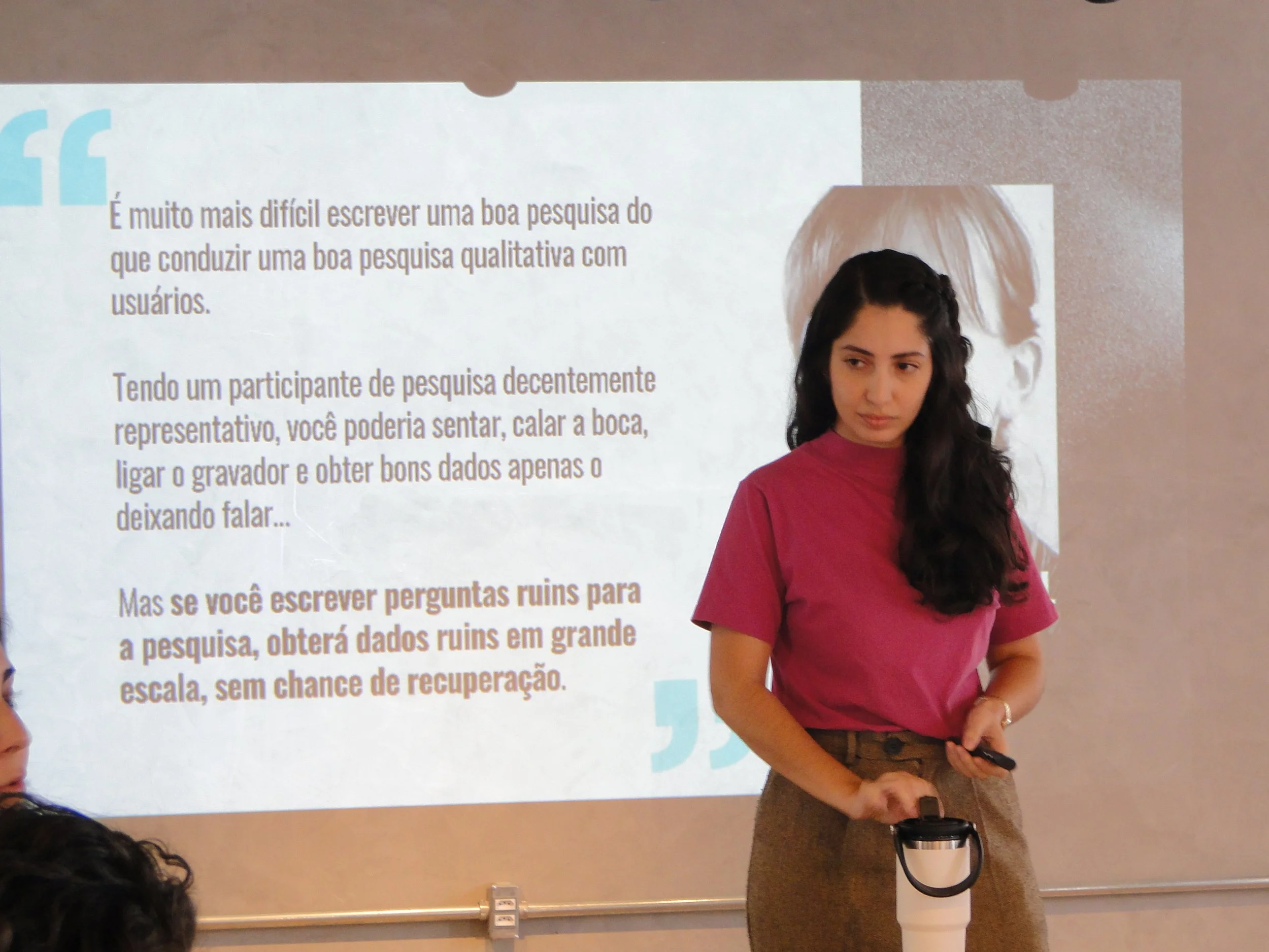 A woman in a pink shirt and brown pants standing in front of a presentation slide with Portuguese text, holding a black remote control and a white thermos.