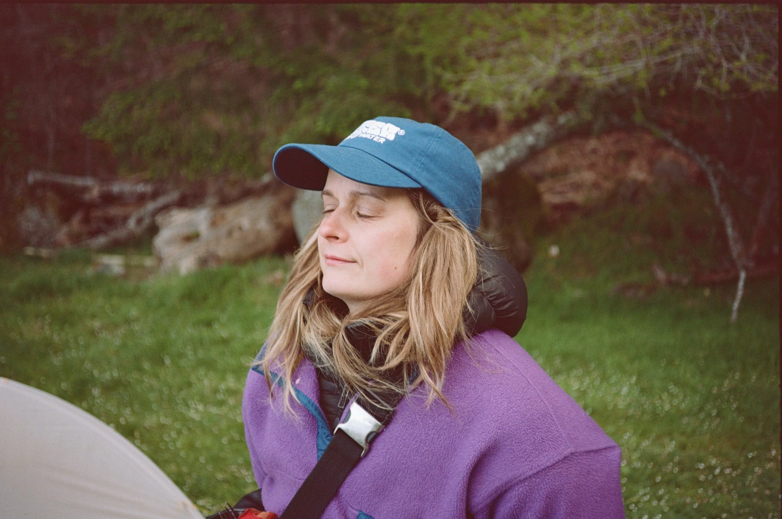 A woman with blonde hair, wearing a blue cap and purple fleece, sits outdoors with her eyes closed, possibly enjoying nature, with trees and grass in the background.