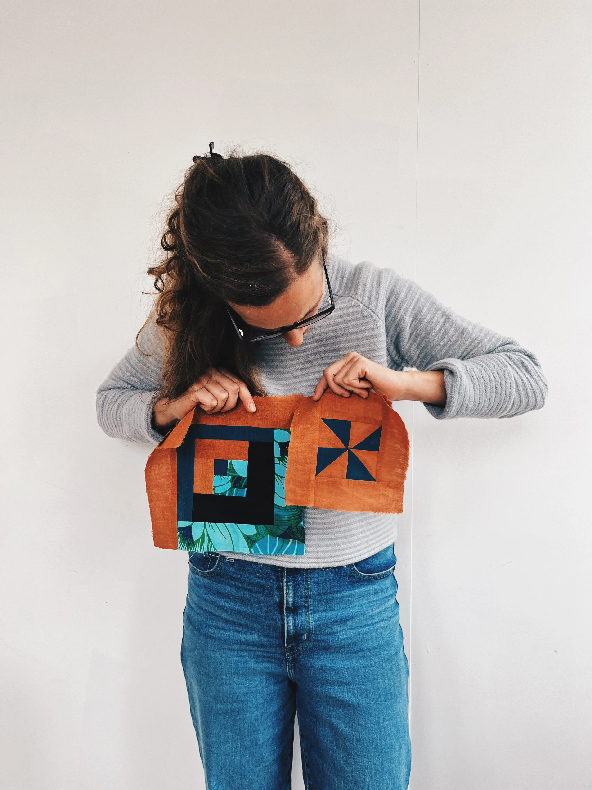 Woman with long brown hair, glasses, wearing a gray sweater and blue jeans, examining a quilt with geometric and floral patterns against a plain white wall.