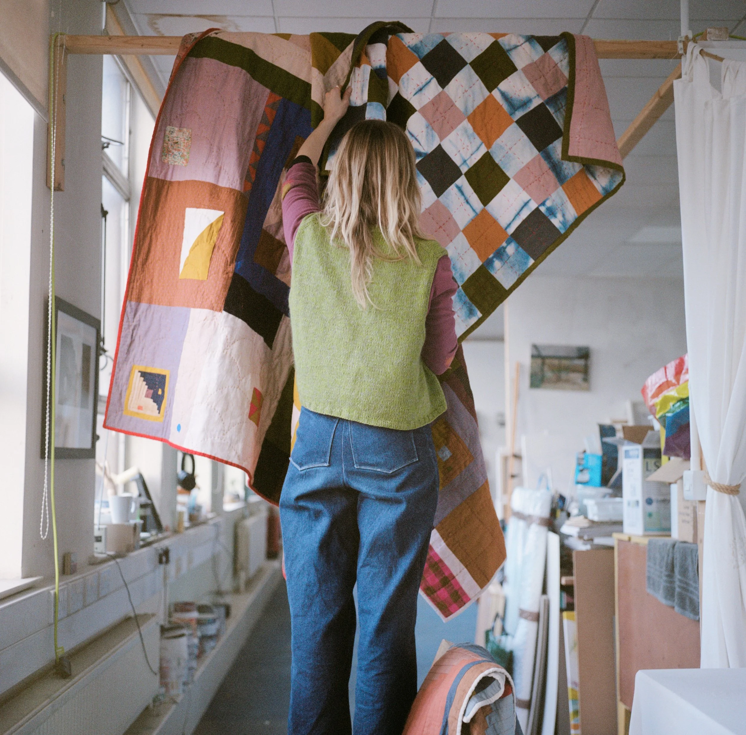 daryl hanging up her quilts in a studio space facing away and wearing blue jeans and a green knitted vest