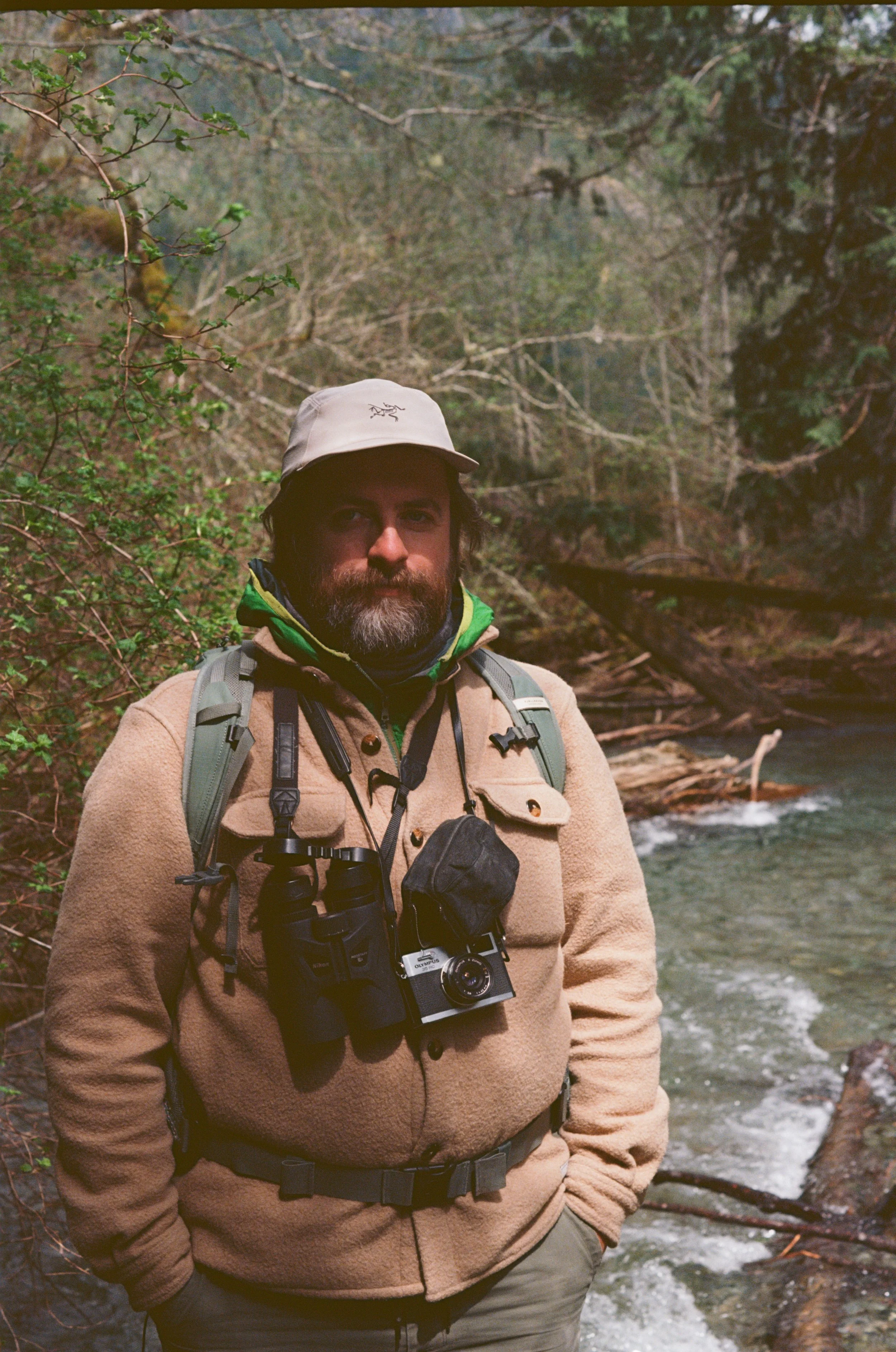 A man with a beard in outdoor gear standing by a stream in a forested area.