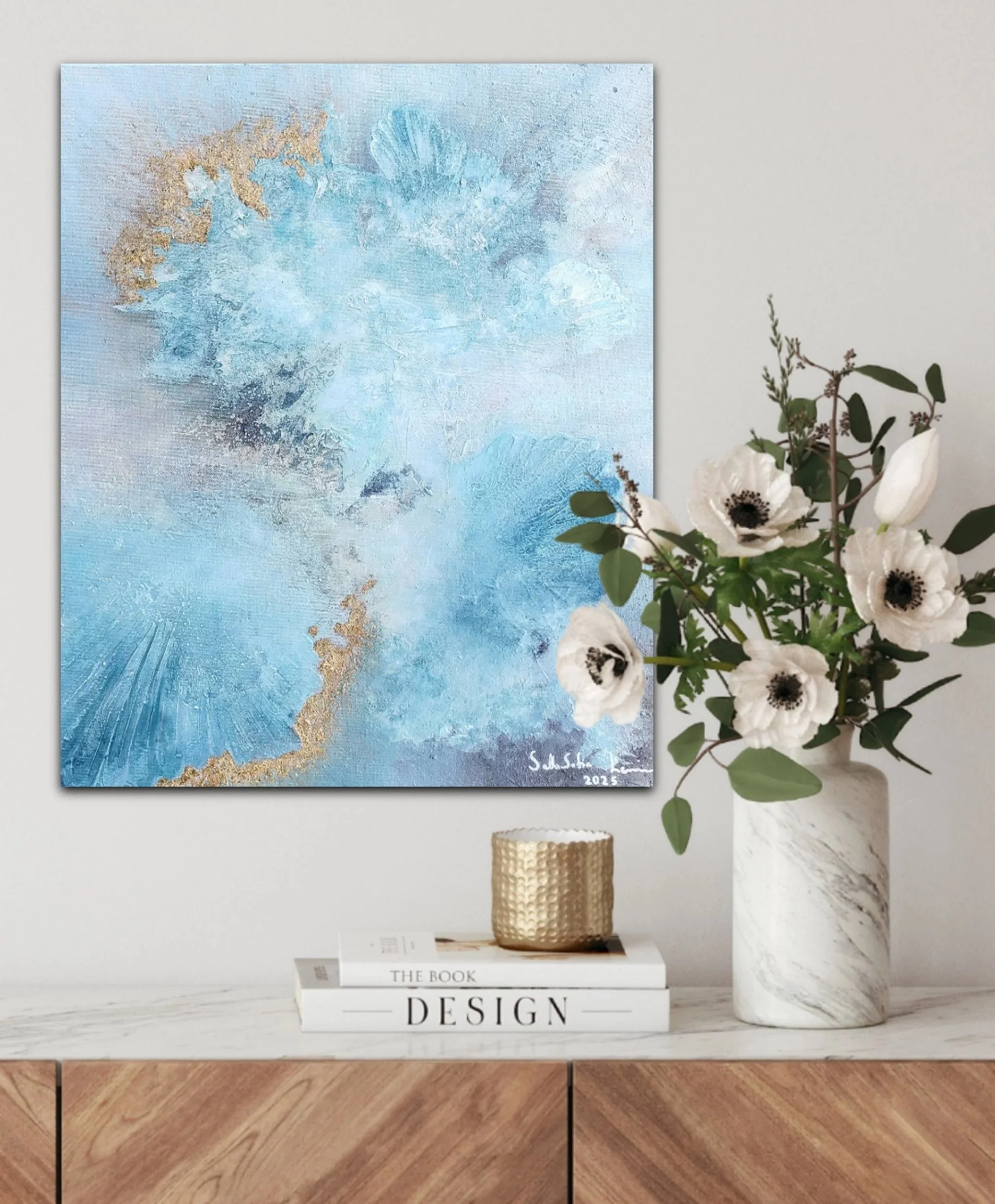 Decorative interior with an abstract blue and gold painting on the wall, a vase with white flowers and greenery, and stacked books on a white marble surface.