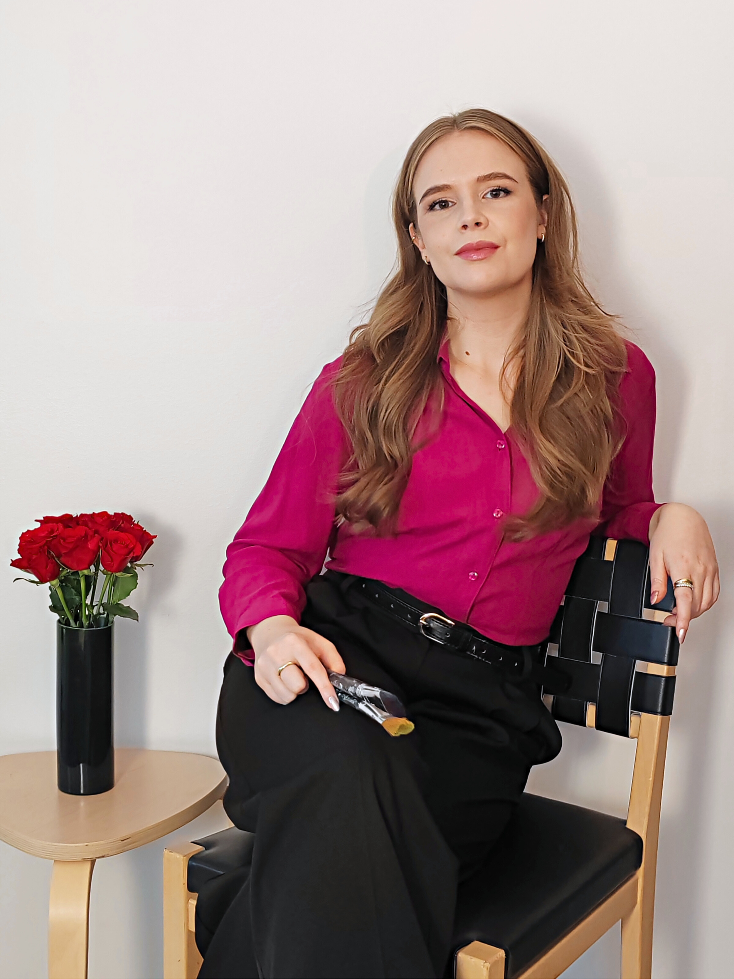 A woman with long, wavy, light brown hair sitting on a black and wood chair against a plain white wall. She is wearing a fuchsia blouse and black pants, holding a paintbrush in her right hand. There is a small wooden side table with a black vase of red roses beside her.