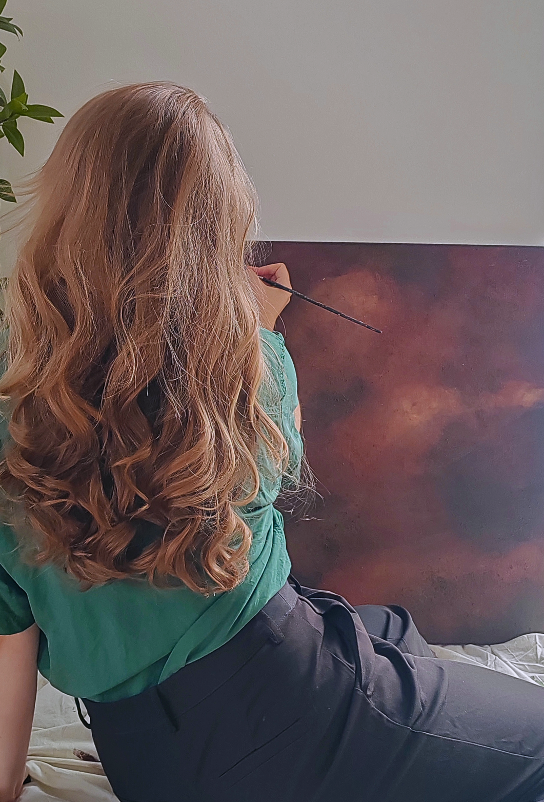 Person with long, wavy hair sitting on a bed, painting a brown and beige canvas with a paintbrush.