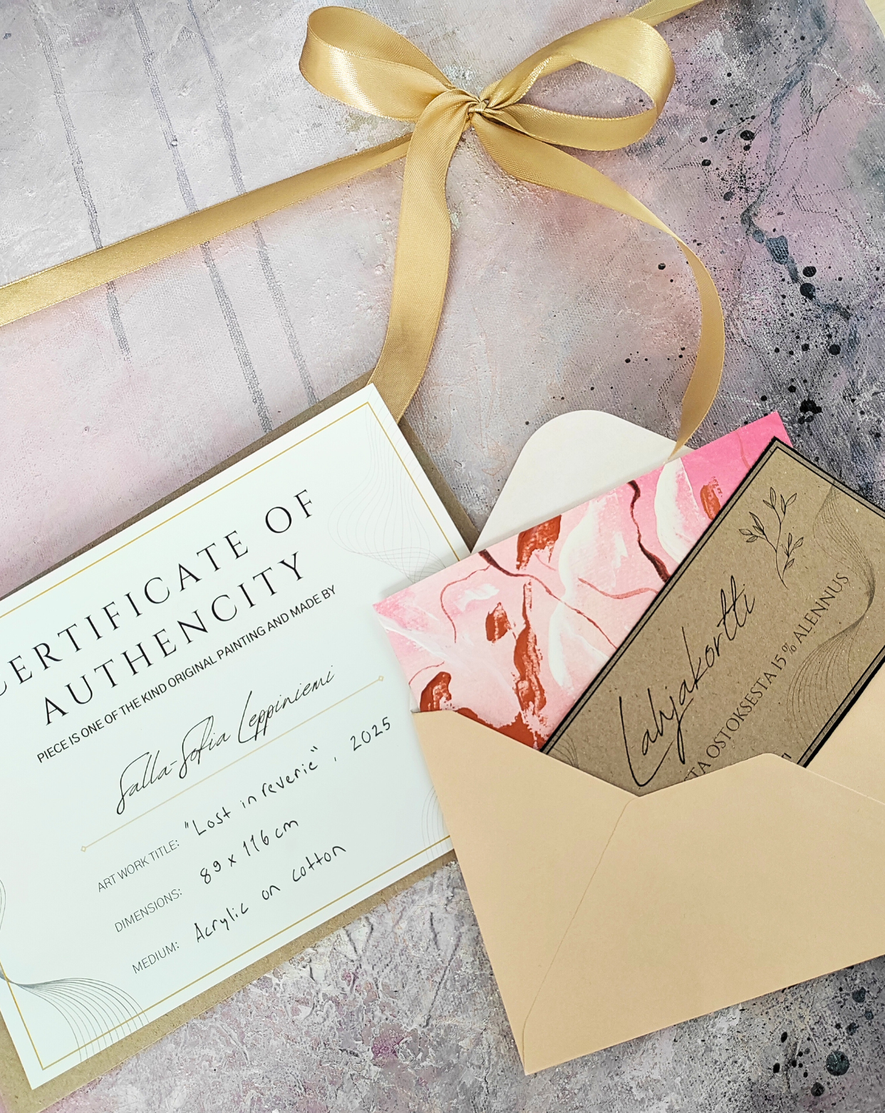 A paper certificate of authenticity, a beige envelope containing a pink floral greeting card, and a voucher, all decorated with a gold ribbon on a textured pink and gray surface.