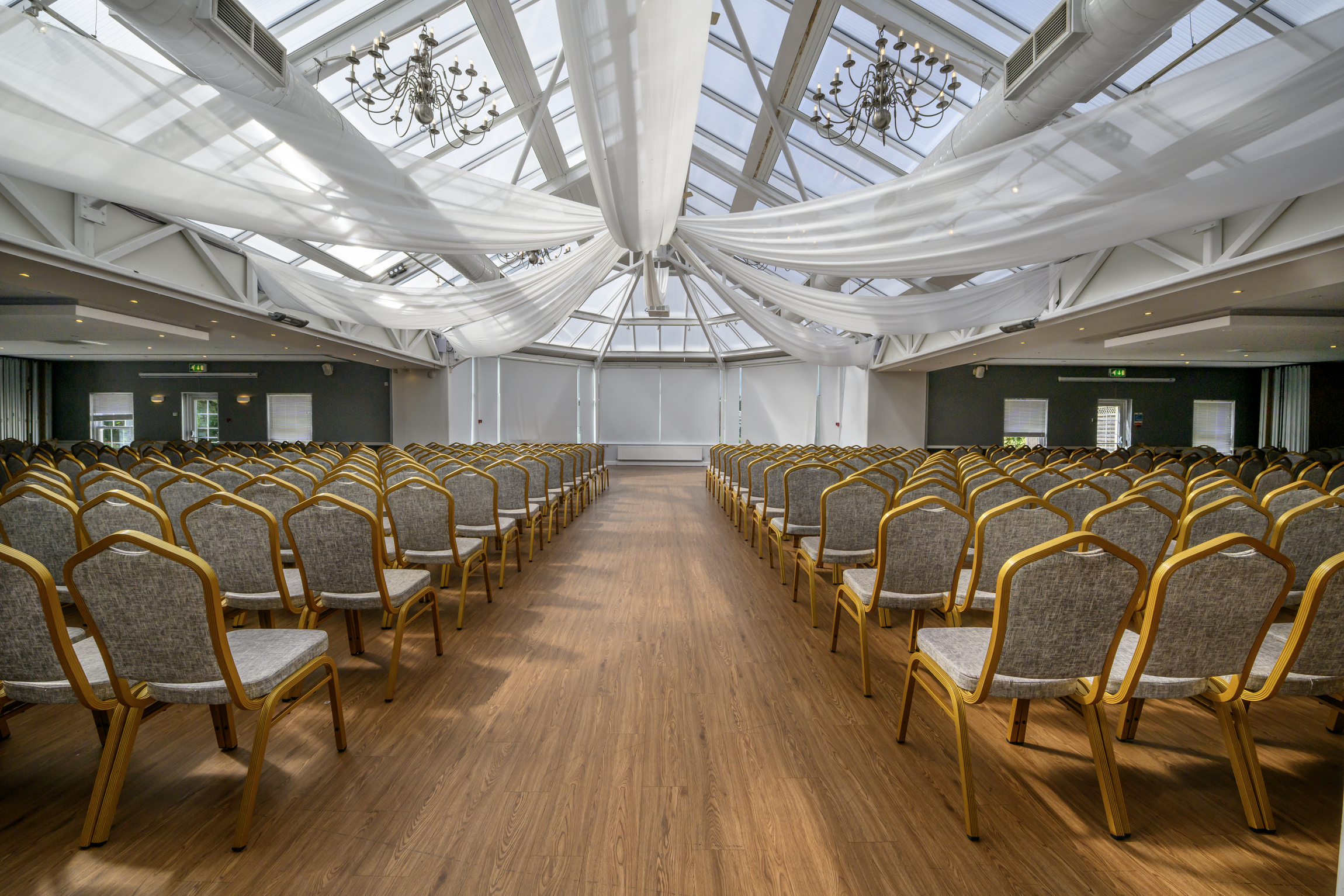 Conference room set up with rows of chairs facing a stage, decorated with white drapes and chandeliers in a glass ceiling venue.