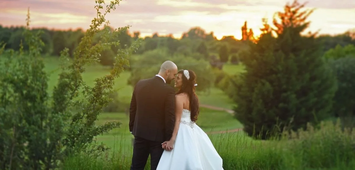 A bride and groom standing in a green outdoor field during sunset, holding hands and touching foreheads, surrounded by trees and rolling hills.