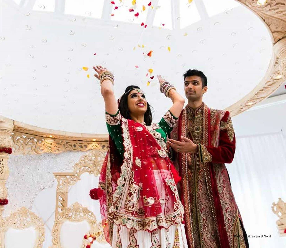 A couple dressed in traditional Indian wedding attire celebrating under a decorated canopy with flower petals falling.