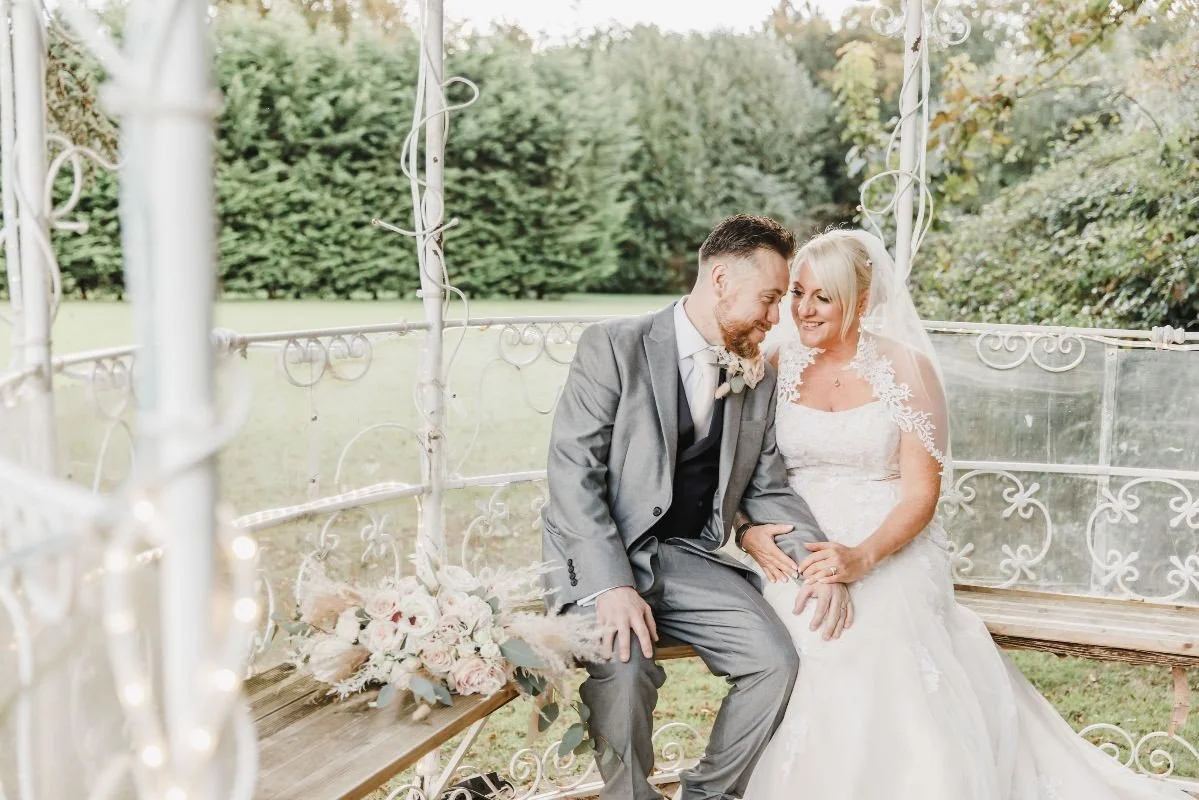 A newlywed couple sits on a vintage wooden swing, smiling and leaning close together outdoors, with lush greenery in the background.