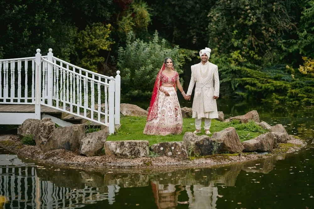 A newlywed Indian couple dressed in traditional wedding attire standing hand-in-hand on a small rocky island with a pond. The bride wears a red and gold lehenga with a red dupatta, and the groom wears a cream-colored sherwani and turban. There is a w