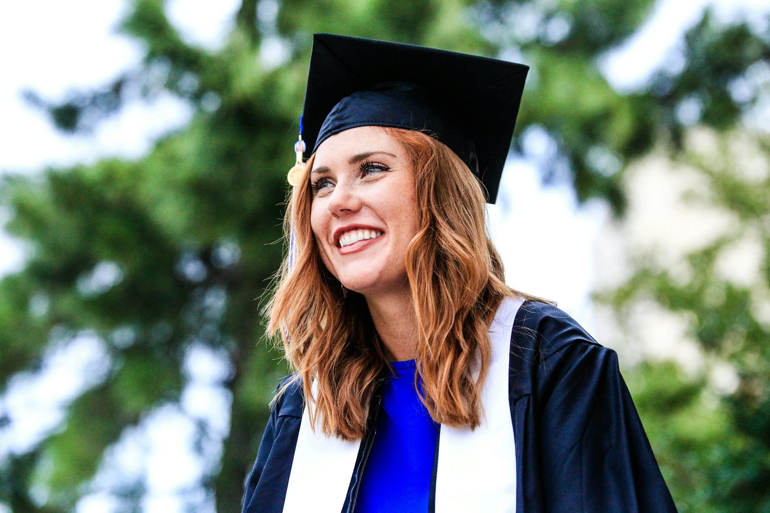A woman in a blue graduation gown and black cap with a tassel smiling outdoors with green trees in the background.