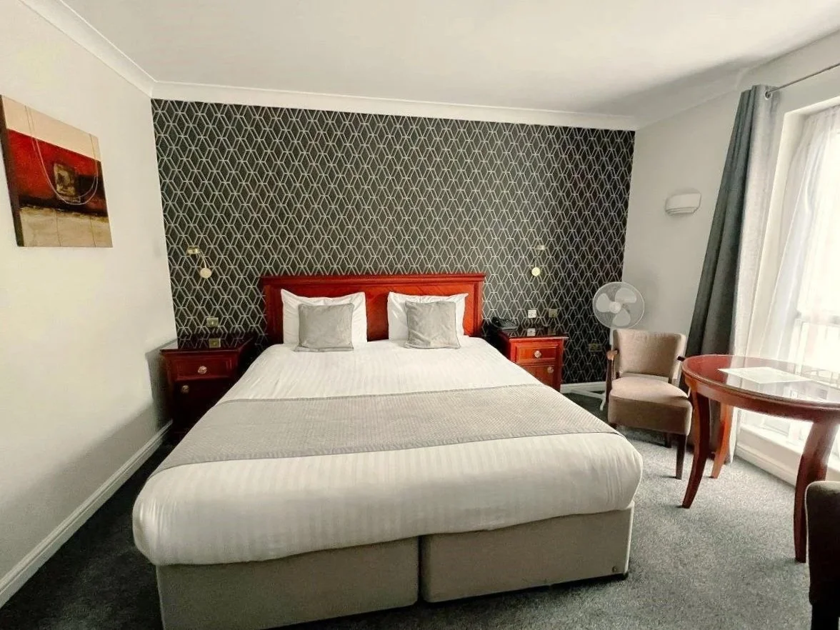 Hotel room with a bed, two nightstands, a table, and chairs, with patterned wallpaper behind the bed and a window with curtains to the right.