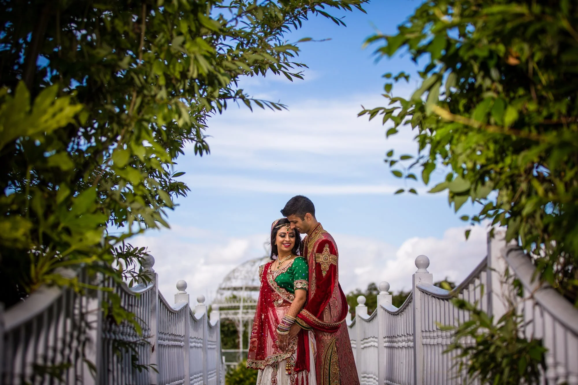 A couple dressed in traditional Indian attire standing close together on a bridge with greenery around and a partly cloudy sky in the background.