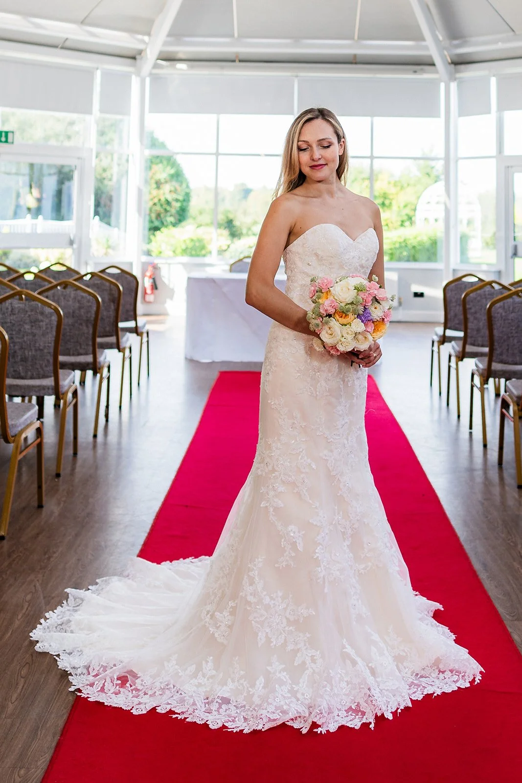 A bride in a strapless lace wedding gown holding a pastel-colored bouquet standing on a red carpet in a bright indoor wedding ceremony space with large windows and empty chairs.