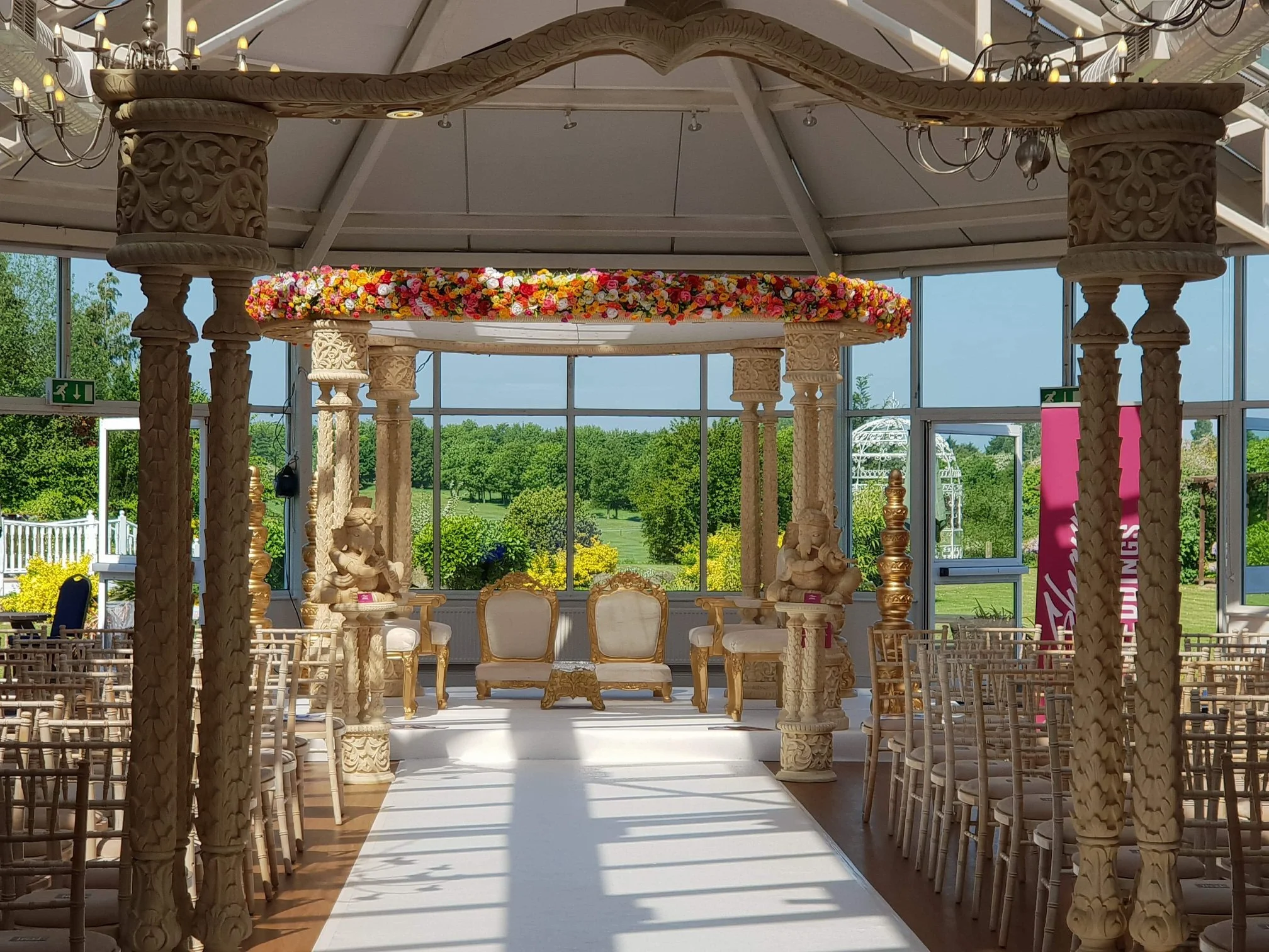 Indoor wedding altar decorated with carved wooden pillars, a floral garland, and seating for the bride and groom, with rows of chairs on either side and large windows overlooking a green outdoor landscape.