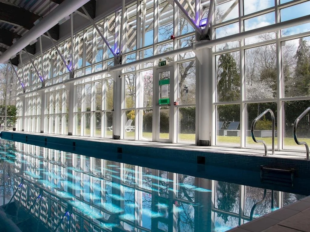 Indoor swimming pool with large glass windows showing outdoor trees and grass, pool ladder on right side, and reflections on the water.