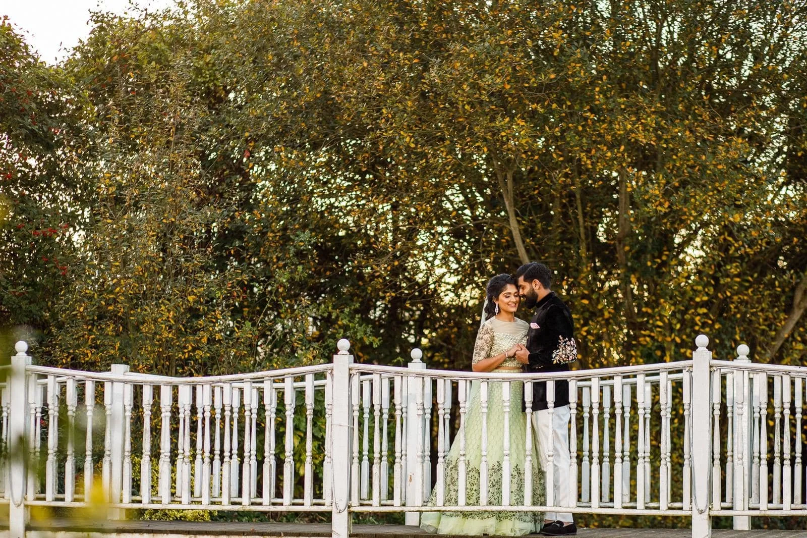 A couple in traditional Indian wedding attire standing on a white fence, smiling and holding hands, with dense green foliage in the background during sunset.