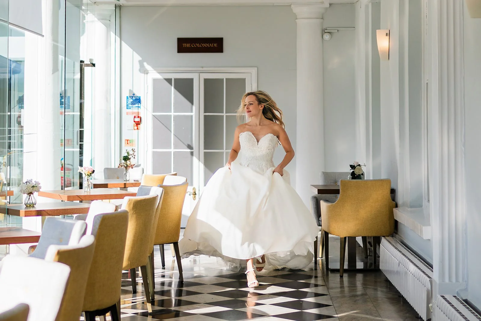 A woman in a white wedding dress walking inside a bright, elegant restaurant or cafe with large windows and checkered floor