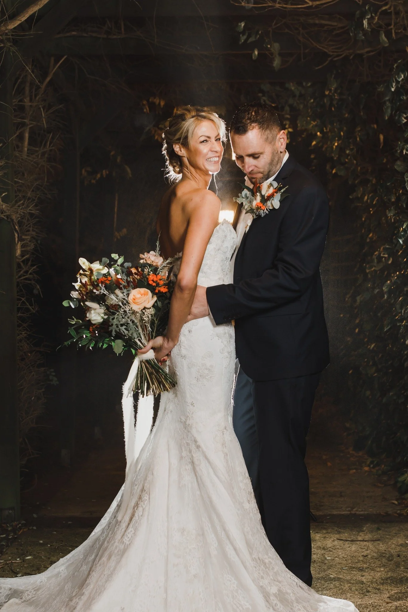 A bride and groom stand together in a night-time outdoor setting, with the bride smiling and holding a bouquet of flowers, and the groom looking down, both dressed in wedding attire.
