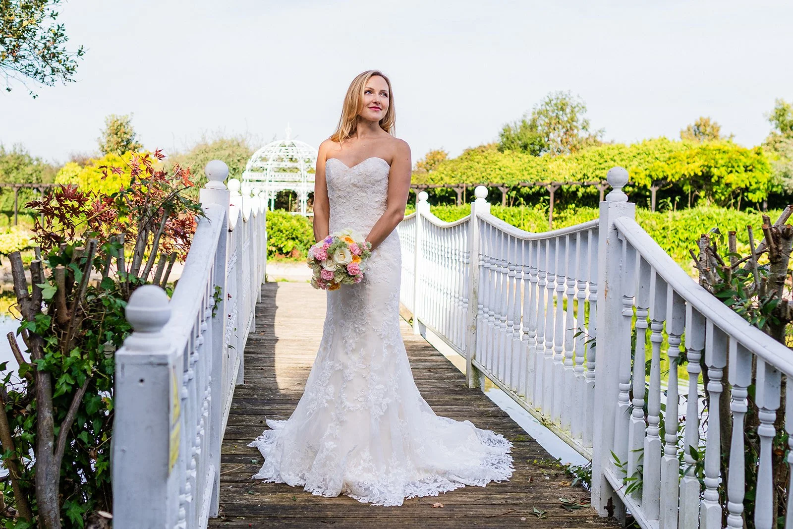 A bride in a white wedding gown holding a bouquet standing on a small wooden bridge outdoors, with greenery and a white gazebo in the background.