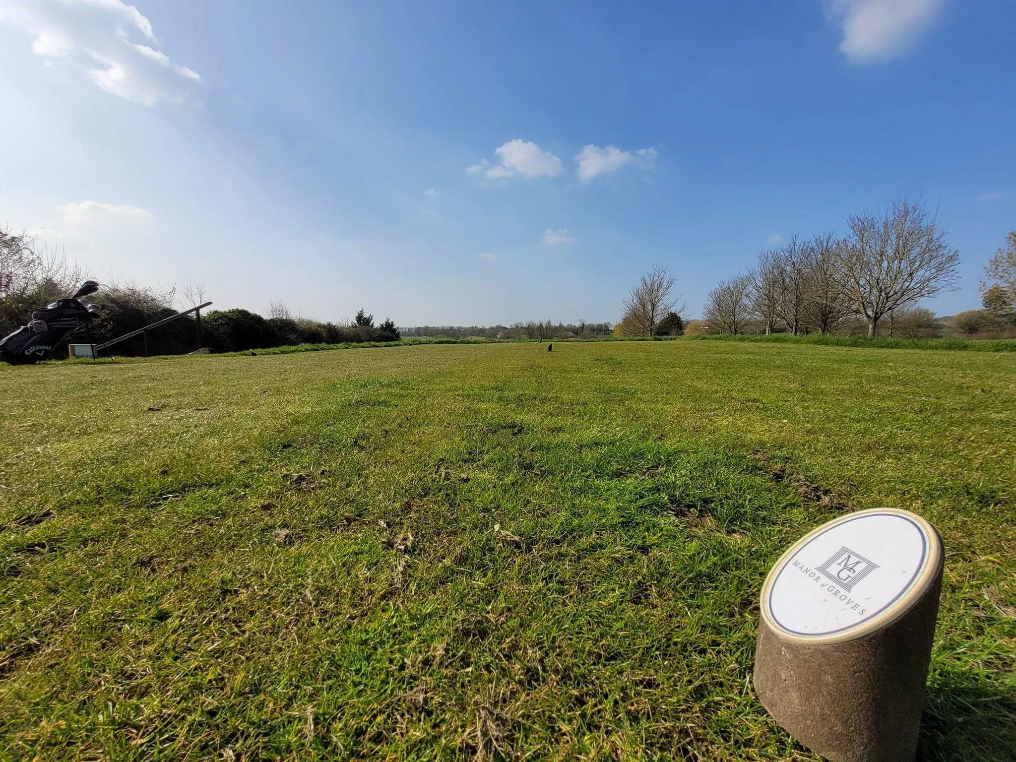 A golf course on a sunny day with a clear blue sky and some clouds. There are trees without leaves in the background, and a golf bag with clubs on a stand on the left side. In the foreground, there is a cylindrical marker with a logo and the text 'MANOR GROVES' on the grass.