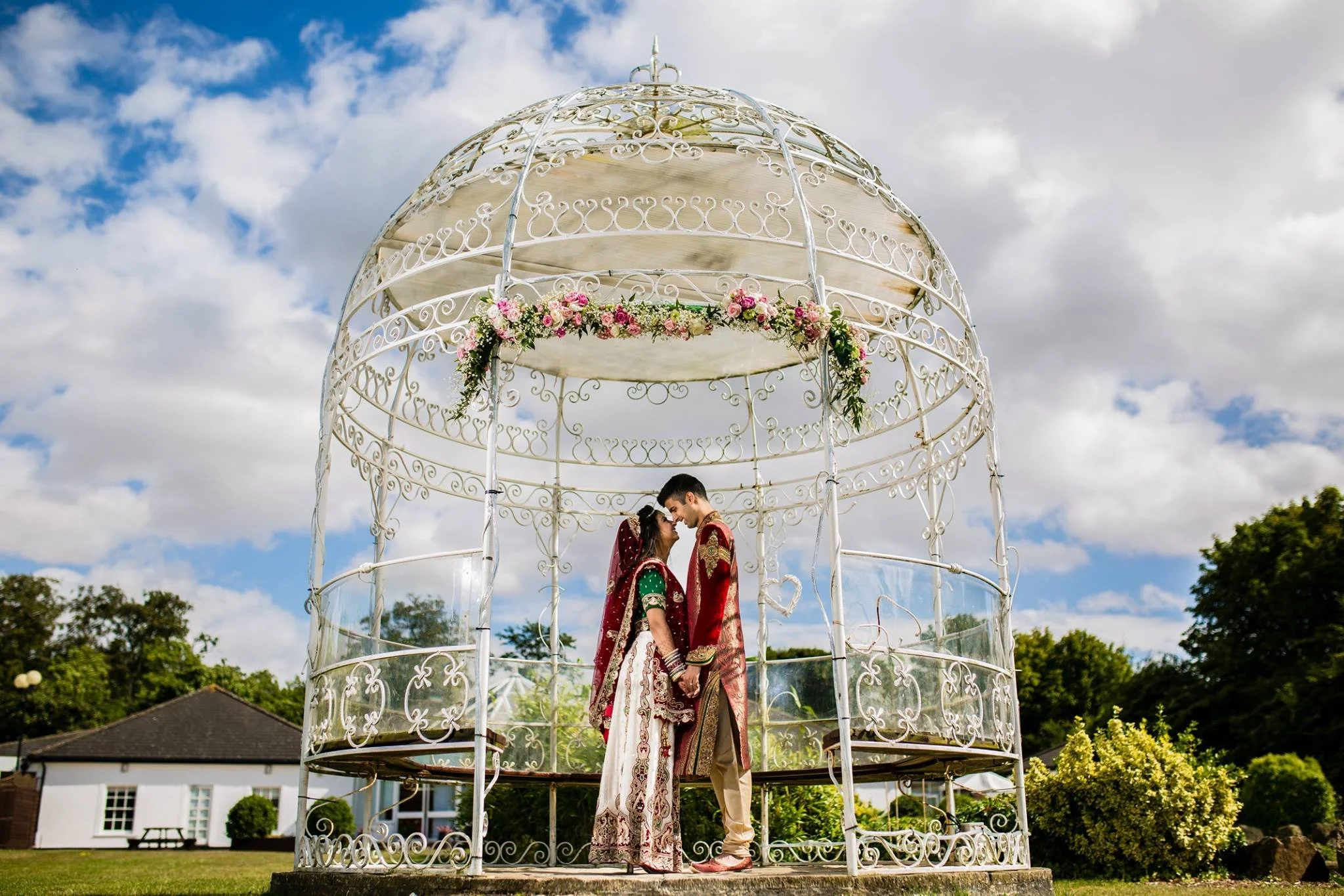 A couple dressed in traditional Indian wedding attire standing close together inside a decorative white gazebo with floral decorations, outdoors on a bright day with blue sky and clouds.