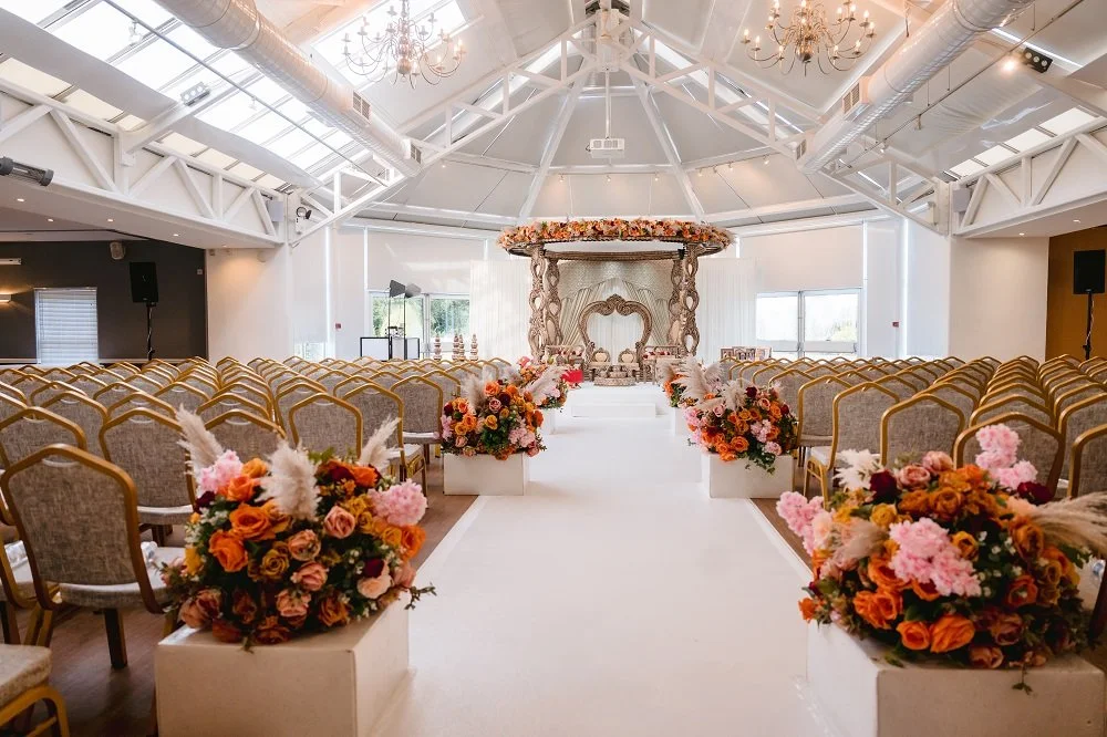 Wedding ceremony setup in a bright, spacious hall with white walls and ceiling, chandeliers, floral arrangements along the aisle, and a decorated altar at the front.