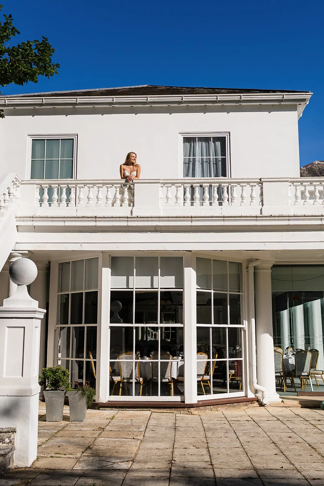 A woman standing on a balcony of a white building, looking to her left against a clear blue sky, with large glass windows and a patio below with chairs and tables.