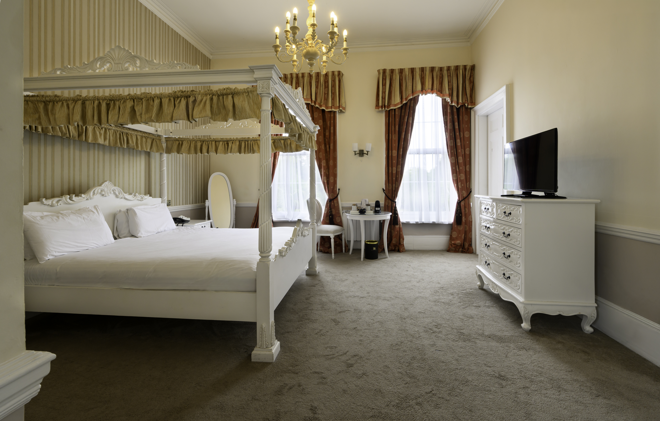 A hotel room with a four-poster bed and ornate bedding, a dresser with a TV, and a seating area near windows with heavy drapes.