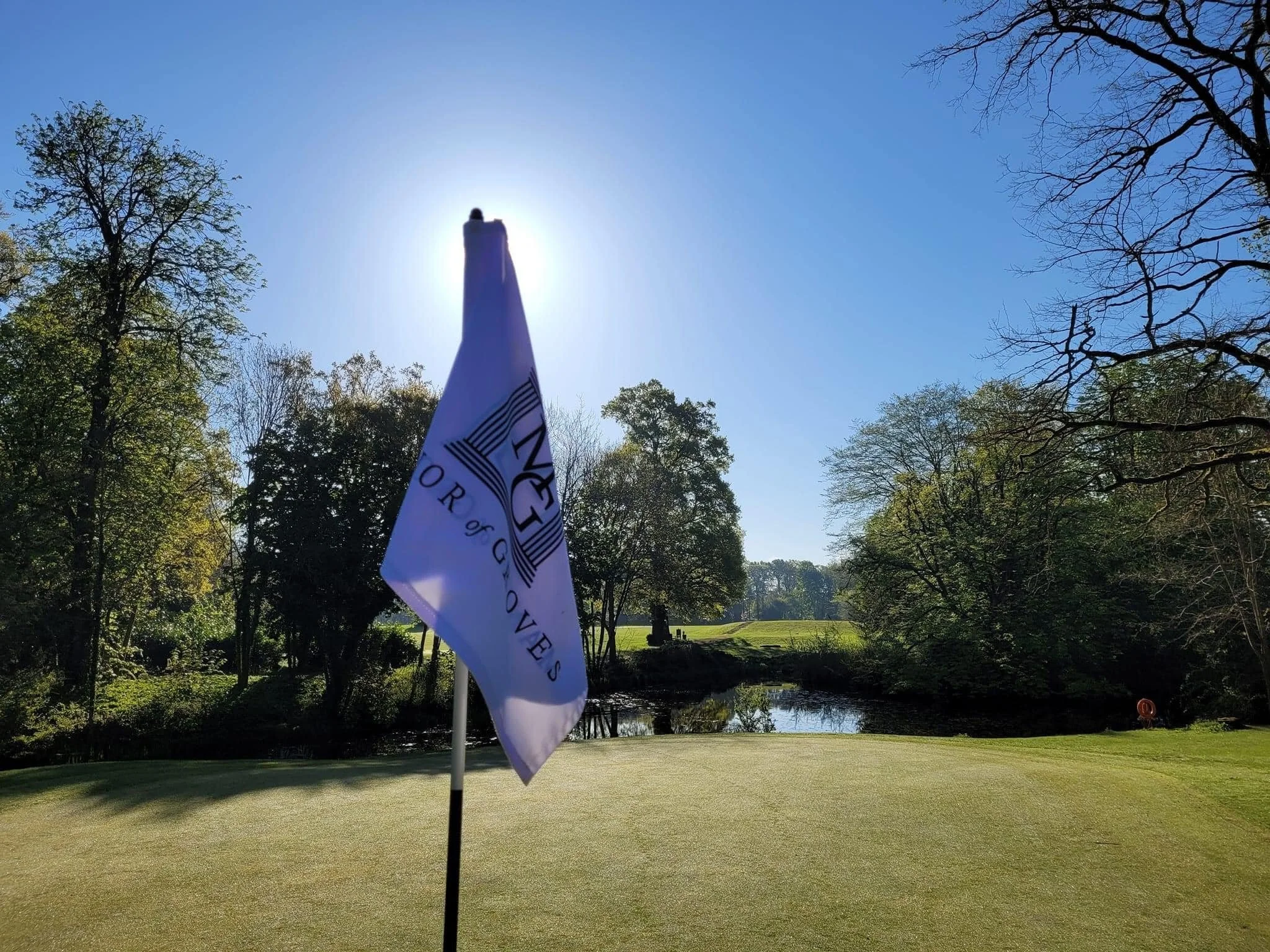 Flag on a golf course green with trees and a pond in the background, sun shining behind the flagpole, blue sky above.