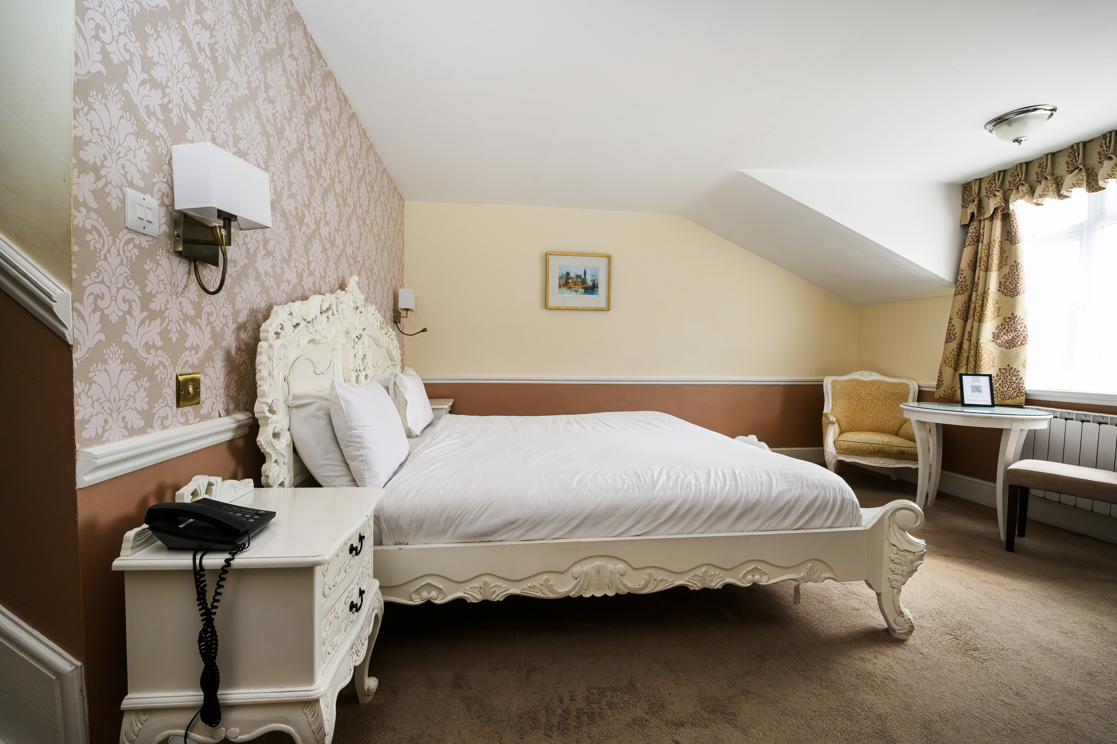 Hotel room with a white ornate bed, a white nightstand with a black telephone, a wall-mounted light fixture, beige and brown wallpaper, a small framed painting, a yellow armchair, a white table with a framed photo, a window with curtains, and a beige