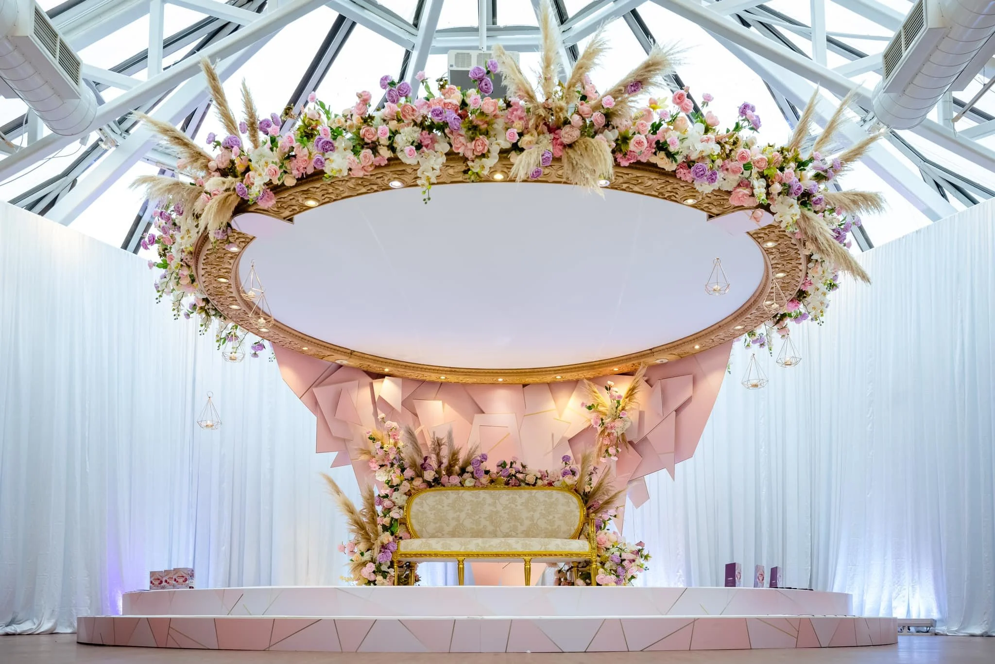 Elegant wedding or event stage with a large round floral arrangement on a gold-trimmed mirror and a decorated throne, with soft pink, purple, and cream flowers, set against white drapes and a glass ceiling with a modern industrial design.