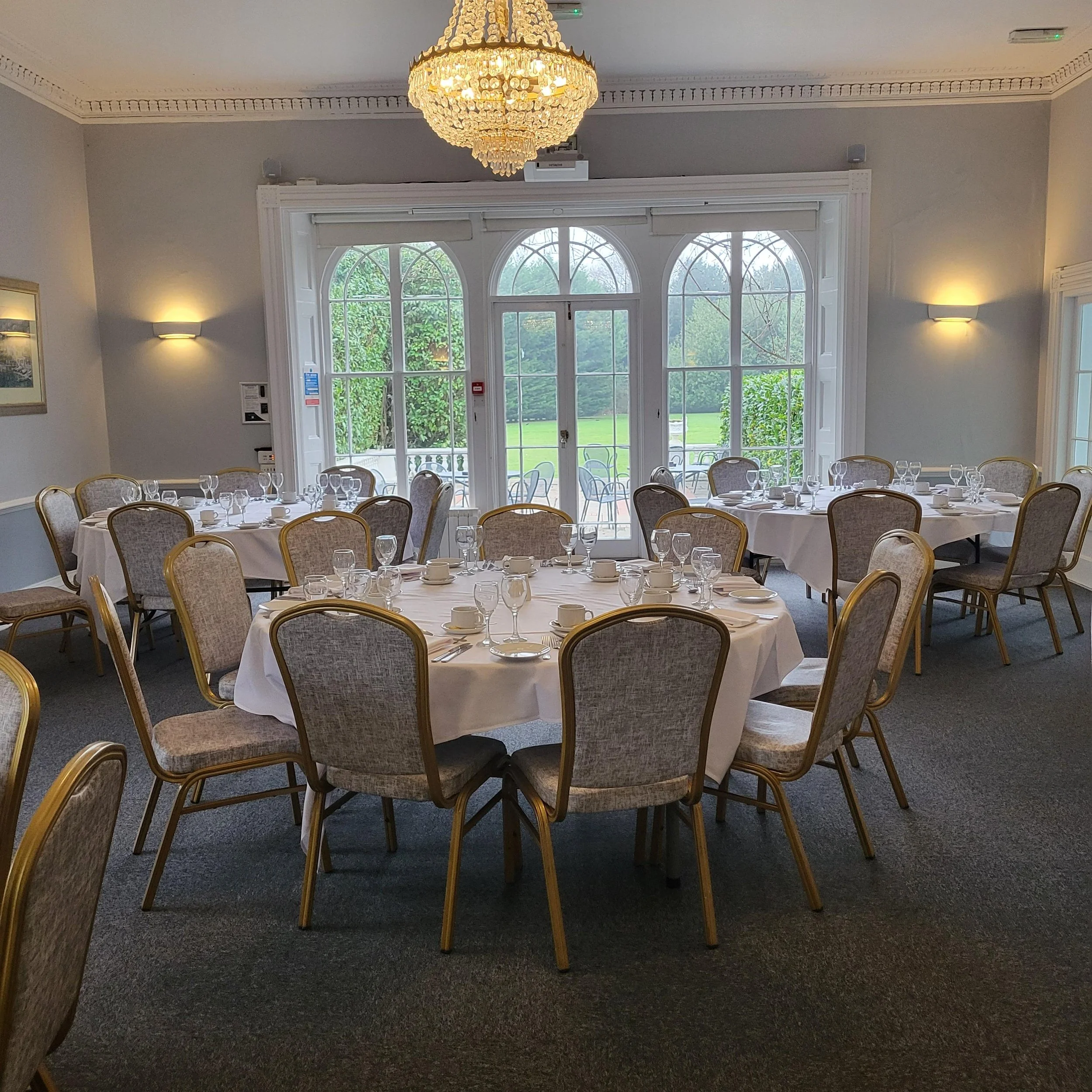 A formal dining room set up with a long table covered with a white tablecloth, set with napkins, glasses, and cups, surrounded by chairs. The room has large windows, red carpet, and a chandelier hanging from the ceiling.