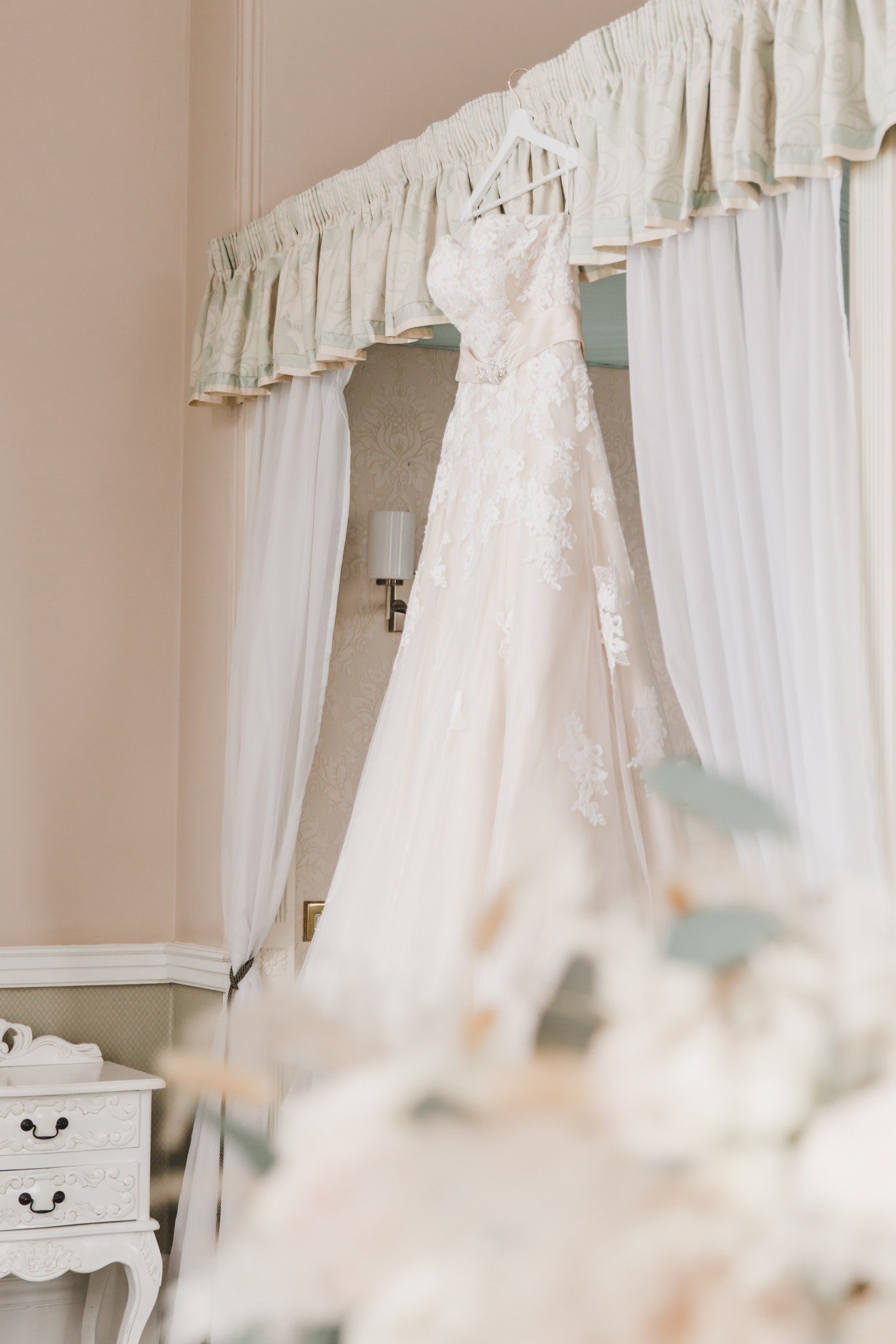 A wedding dress hanging from a white hanger on a canopy bed with white curtains in a beige-colored room.