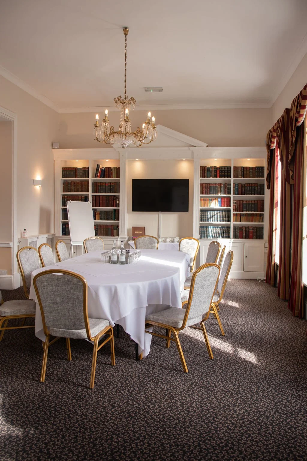 A formal dining room with round tables covered in white tablecloths, set with white napkins, cups, and glassware. There is a large bookshelf with books and a TV mounted on the wall in the background. The room has a chandelier, striped curtains, and a fireplace with a painting above it.