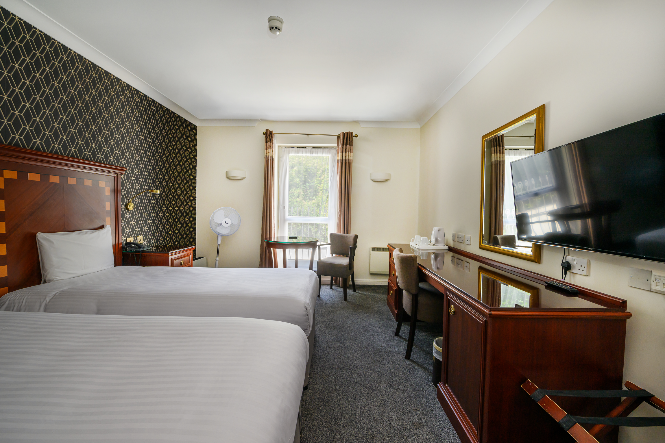 A hotel room with two beds, a flat-screen TV on the wall, a mirror, a small table with a chair, a fan, and window with curtains.