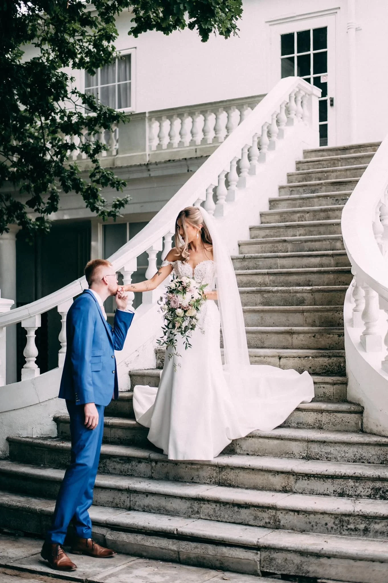 A bride in a white wedding gown and a groom in a blue suit on stairs outside a white building. The groom is kissing the bride's hand, and she is holding a bouquet of flowers.