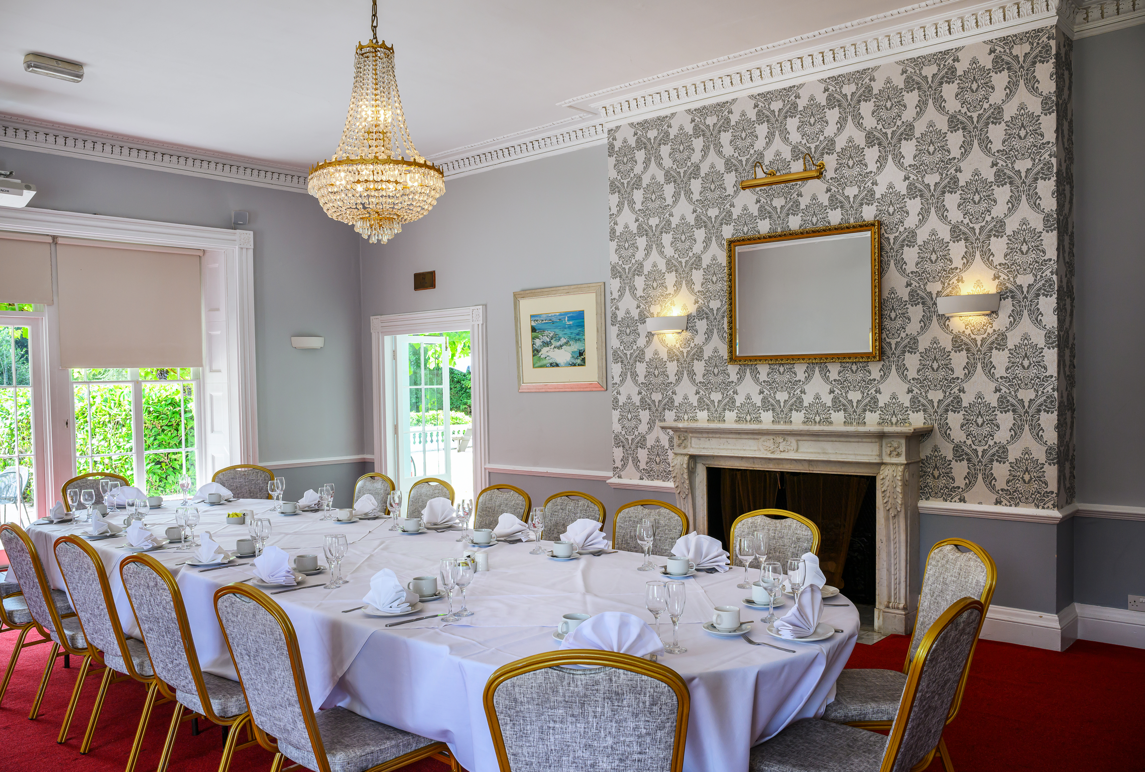 A formal dining room with a large rectangular table set with white tablecloth, napkins, cups, and glasses. The room has a chandelier, a fireplace, patterned wallpaper, and a painting on the wall. Natural light comes through windows and a glass door.