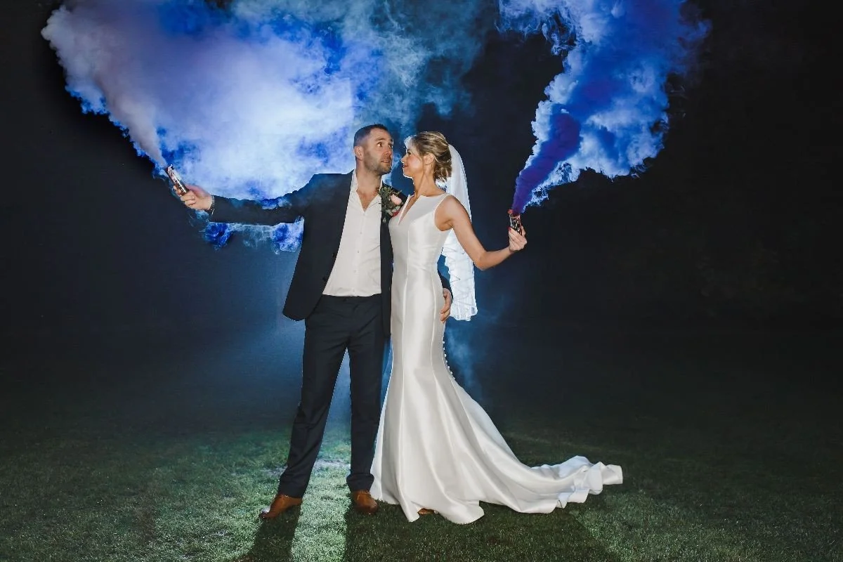 A bride and groom in wedding attire holding blue smoke bombs, standing closely together at night with a dark sky background.