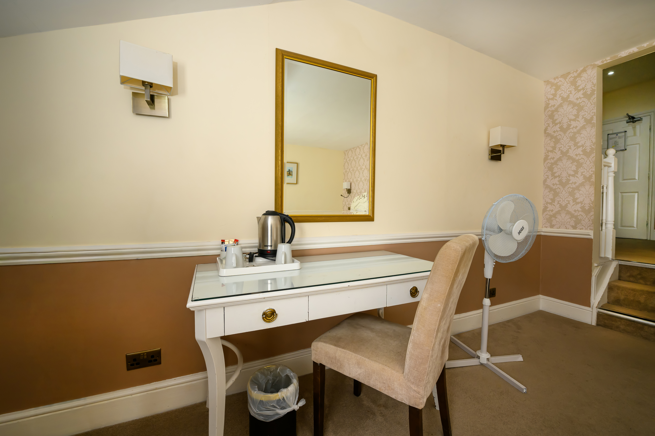 A room with a cream-colored wall and a brown lower wall. There's a white desk with a glass top, a beige upholstered chair, a trash can, a standing fan, a mirror, a wall-mounted light, and a door staircase leading to another room.