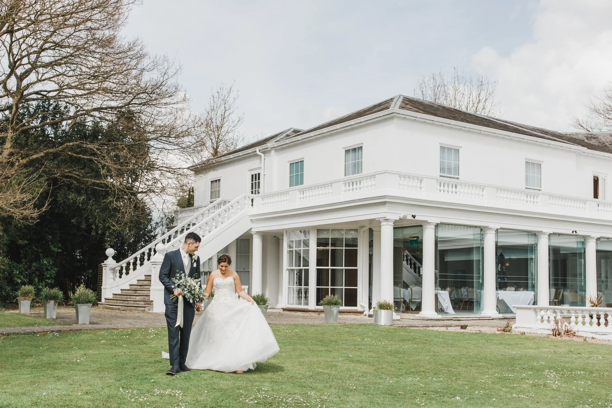 A bride and groom walk hand in hand on a grassy lawn in front of a large white wedding venue with columns and stained glass windows, surrounded by leafless trees.