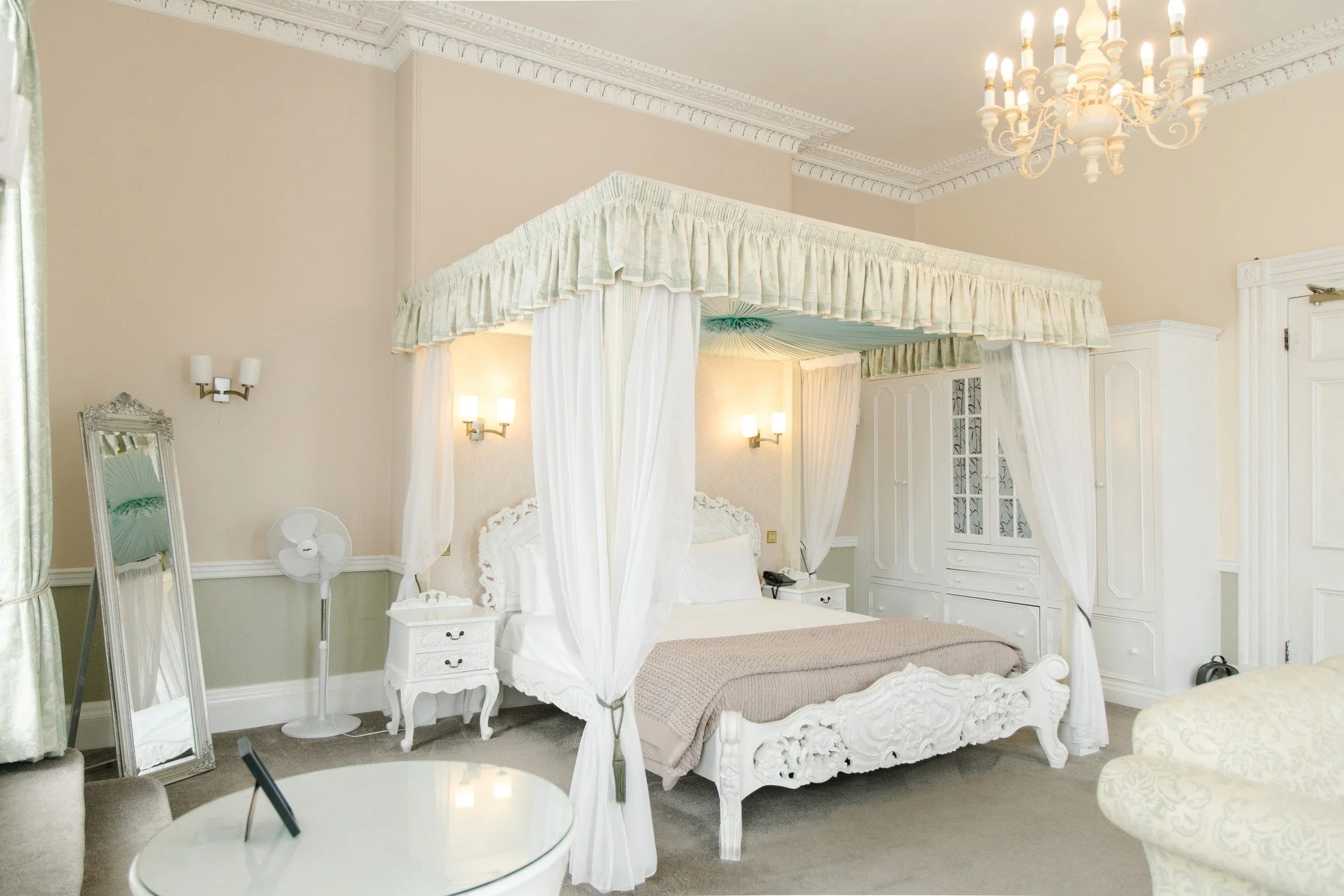 Elegant hotel room with a white four-poster bed, ornate furniture, wall-mounted lamps, a standing mirror, and a chandelier.