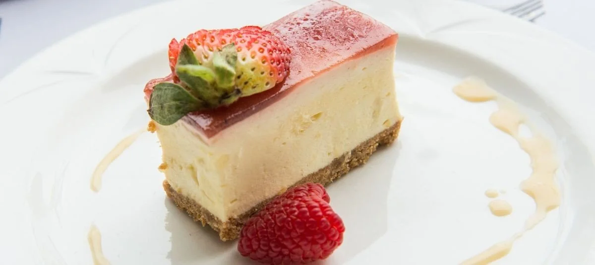A slice of cheesecake topped with a strawberry and a small strawberry on the side, garnished with a light drizzle of sauce on a white plate.