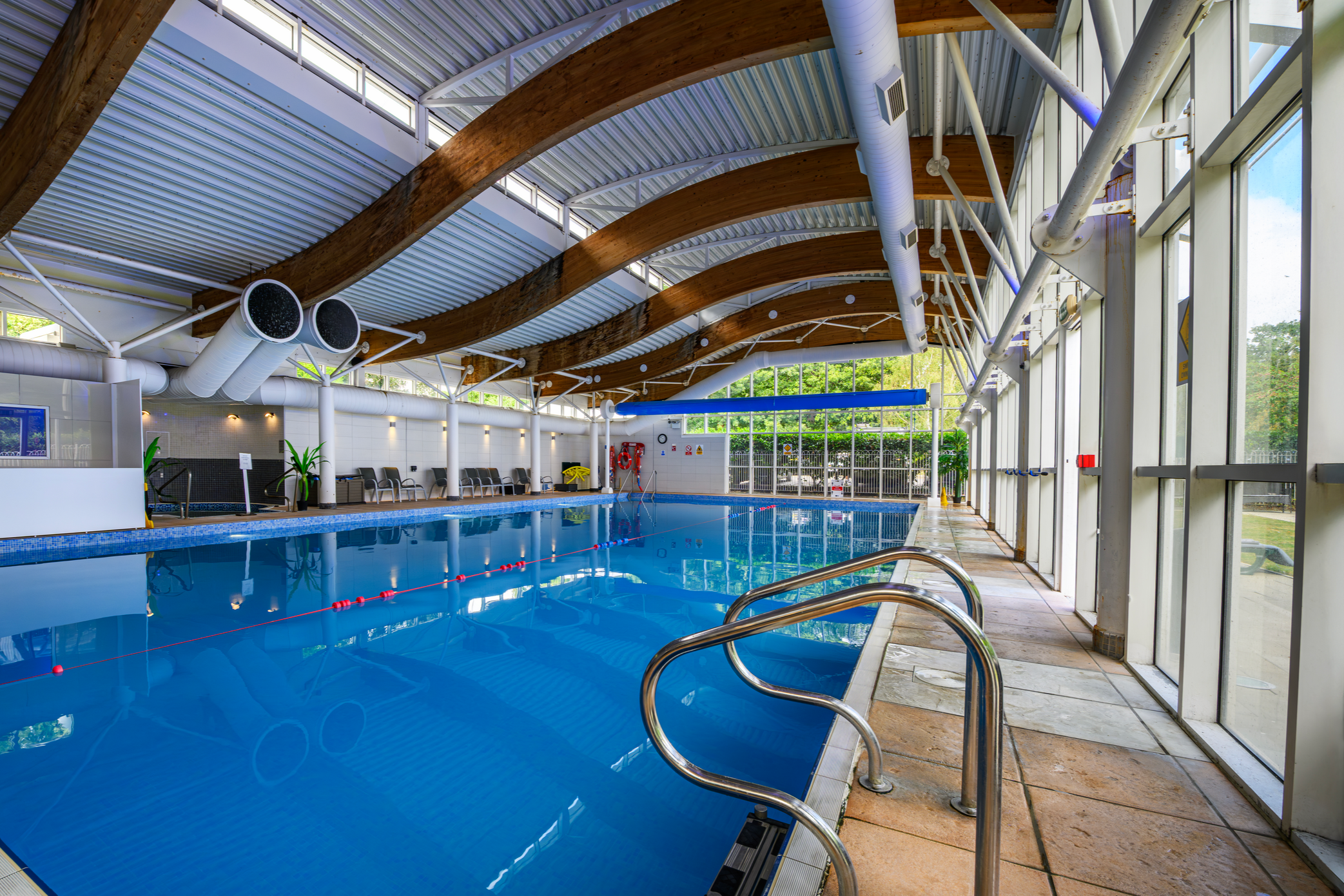 Indoor swimming pool with blue water, glass walls, and wooden beams on the ceiling, surrounded by chairs and safety equipment.