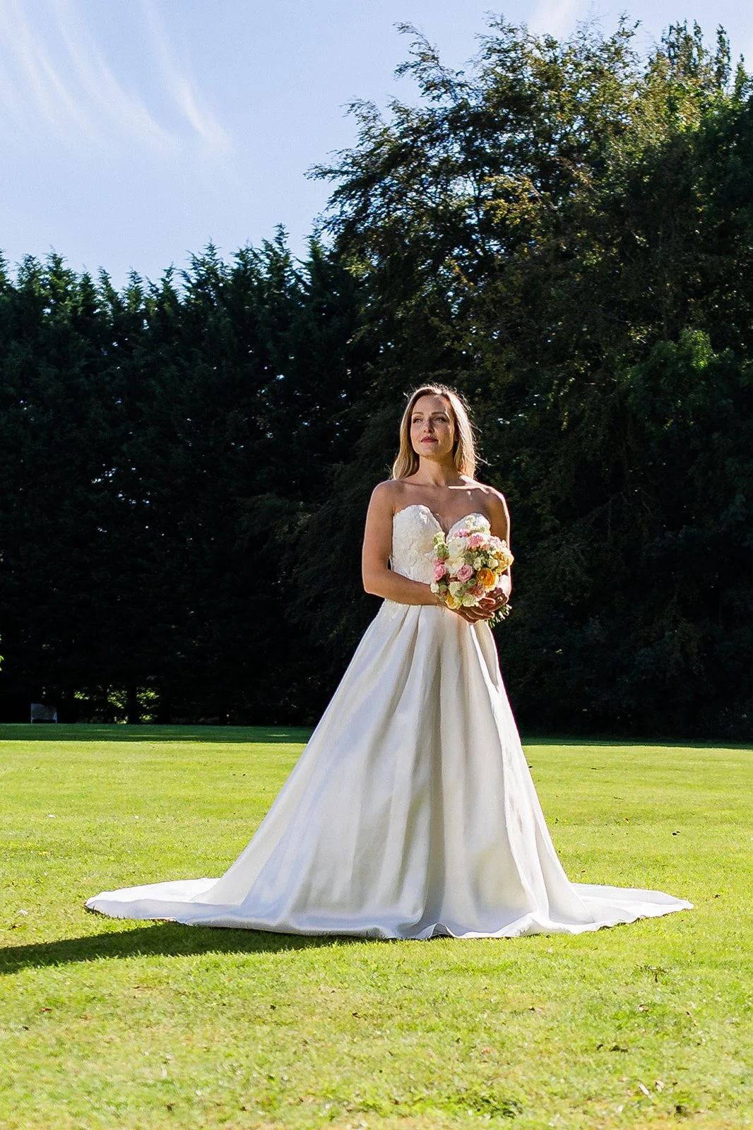 A woman in a white wedding dress holding a bouquet of pink and white flowers outdoors on a sunny day with green trees in the background.