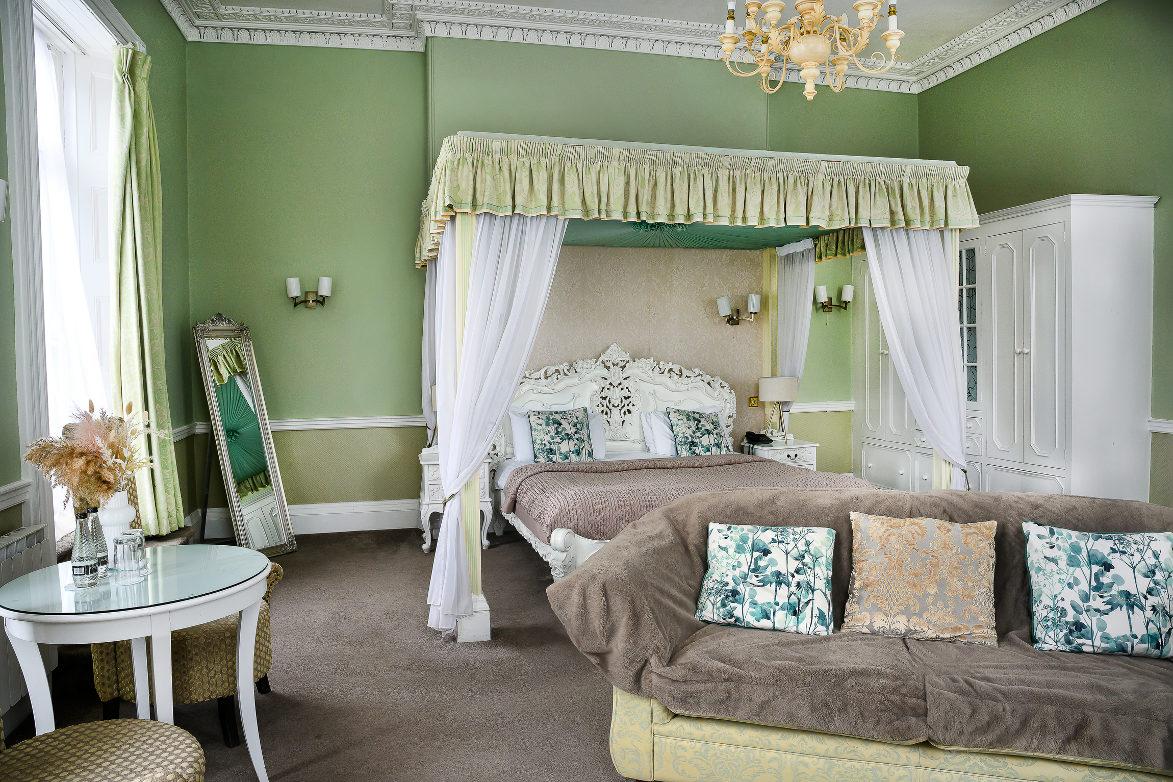 A luxurious bedroom with a four-poster bed with white carved wood, canopy curtains, and cushions. The room has green walls, ornate crown molding, a chandelier, a white wardrobe, and a seating area with a table and chairs.