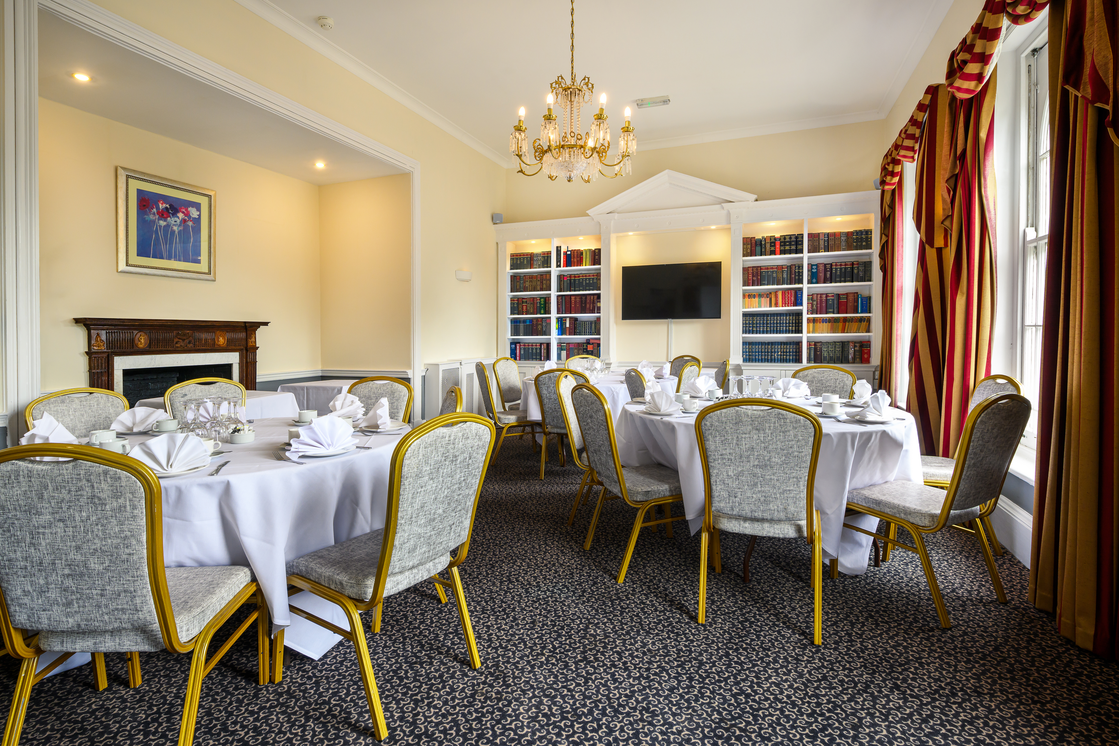 Elegant dining room with round tables covered in white tablecloths, set with napkins, cups, and silverware, yellow-rimmed chairs, a chandelier, a fireplace with a painting above, and large windows with red and gold striped curtains.